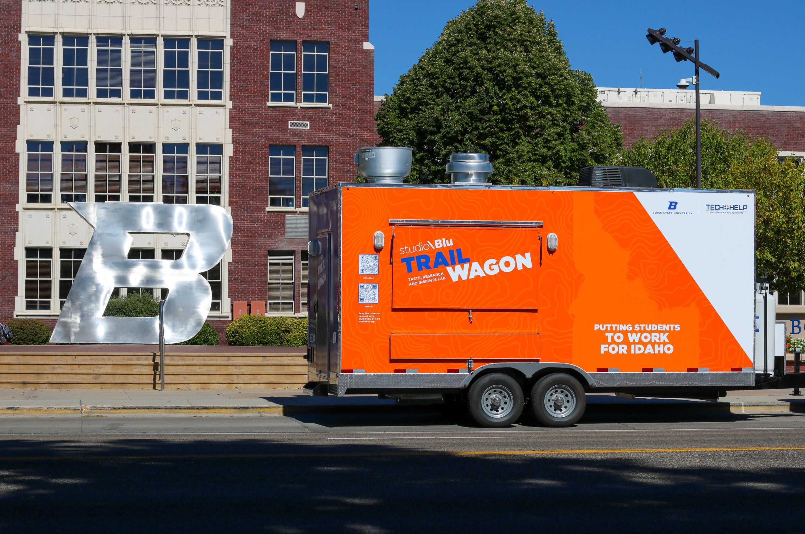 A bright orange food truck trailer that says "TRAIL Wagon" on the side is parked on Boise State's campus in front of the large B.