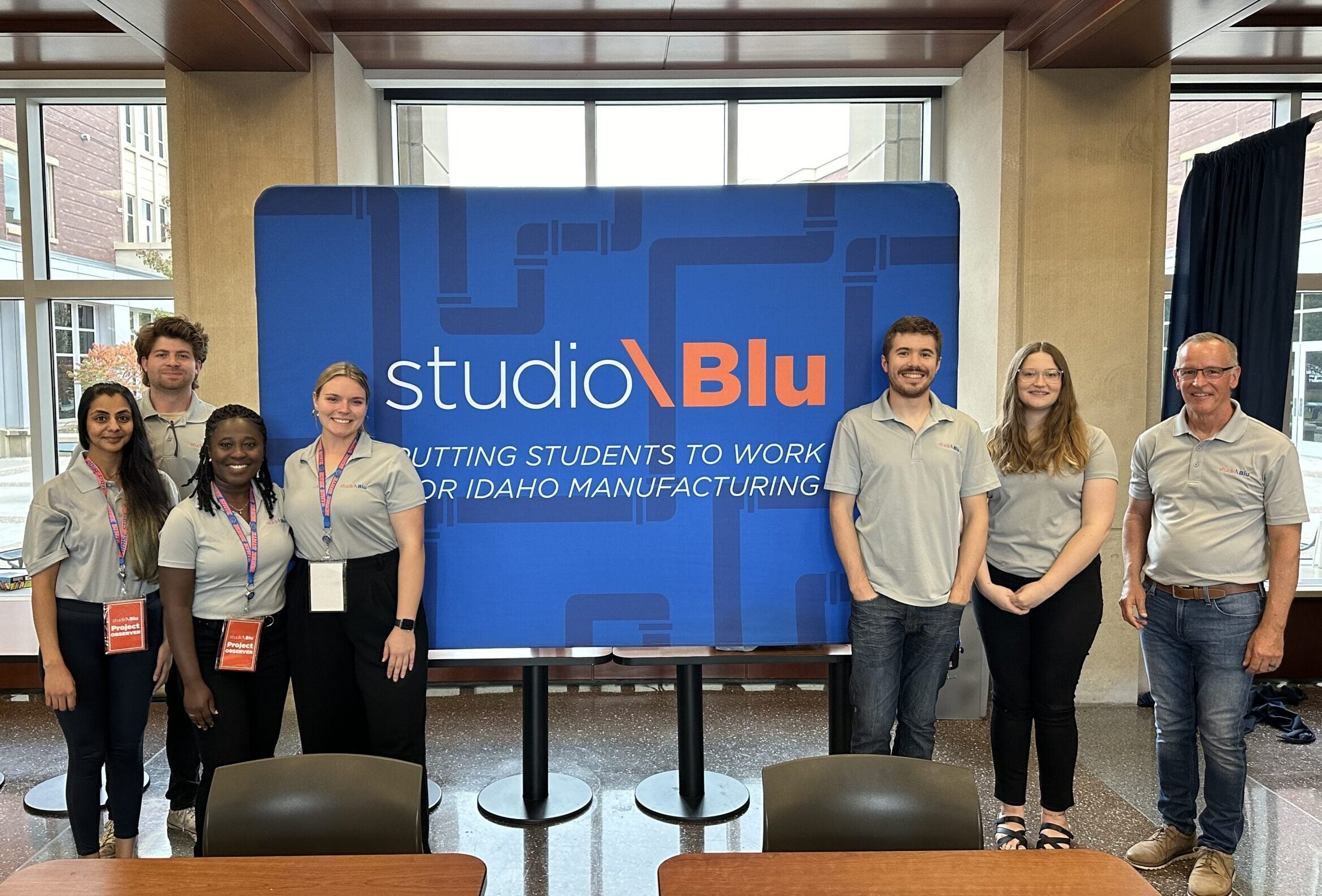 A group of students and their team manager stand in front of a studio\Blu branded backdrop.