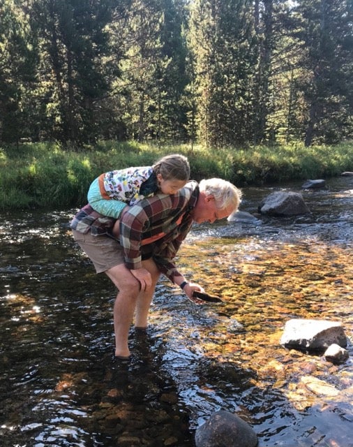 A man looks in the river with a small child on his back. 