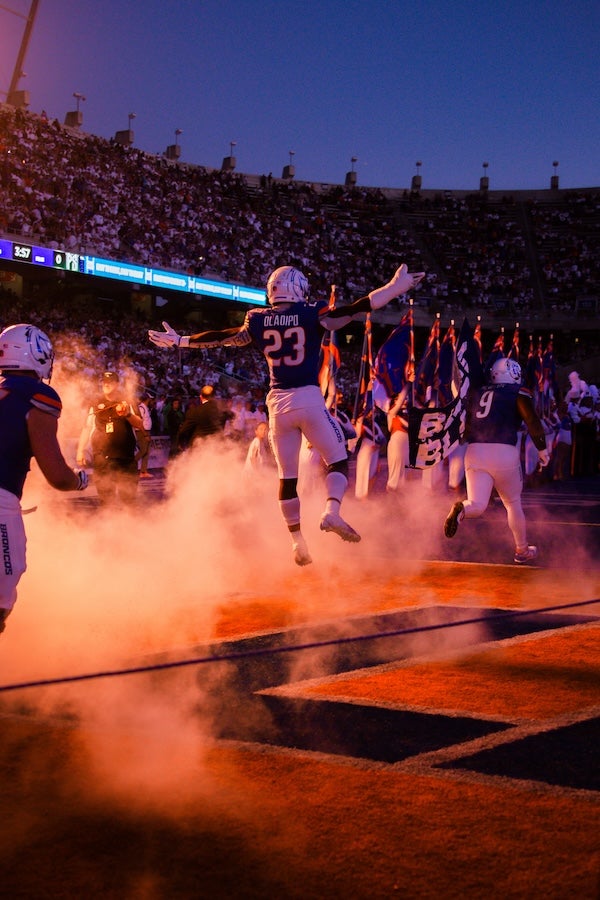 A football player is captured jumping with arms to their side as the team enters the field to blue and orange lights and fog