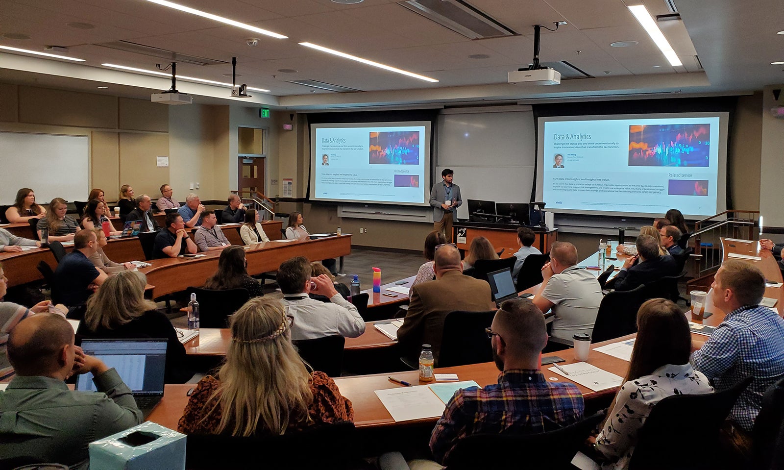 A group of adults sits in a classroom focused intently on a speaker with slides about data and analytics on the screen behind him.