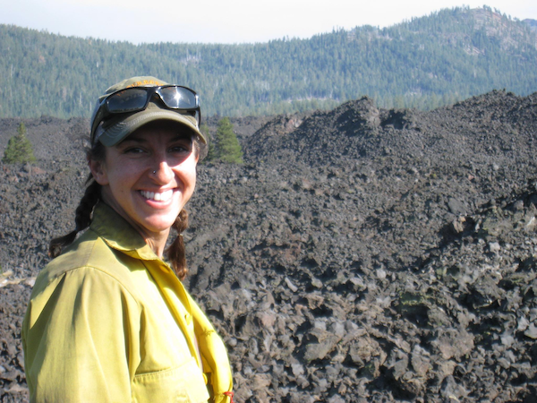 young woman in ball cap and yellow shirt smiles at camera, standing in front of rough, mountainous ground