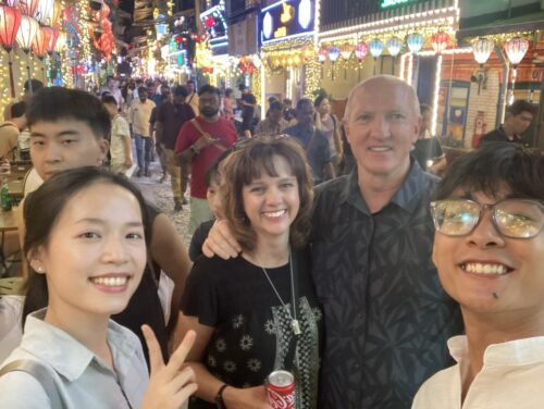 A small group stands in a colorful street market and smiles for the camera.