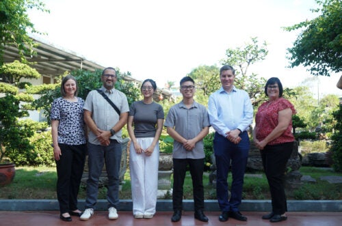 A group of adults and students stand in a line in front of scenic greenery.