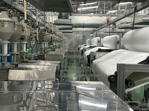 Image of the inside of a factory with rolls of fabric-like material on manufacturing equipment.