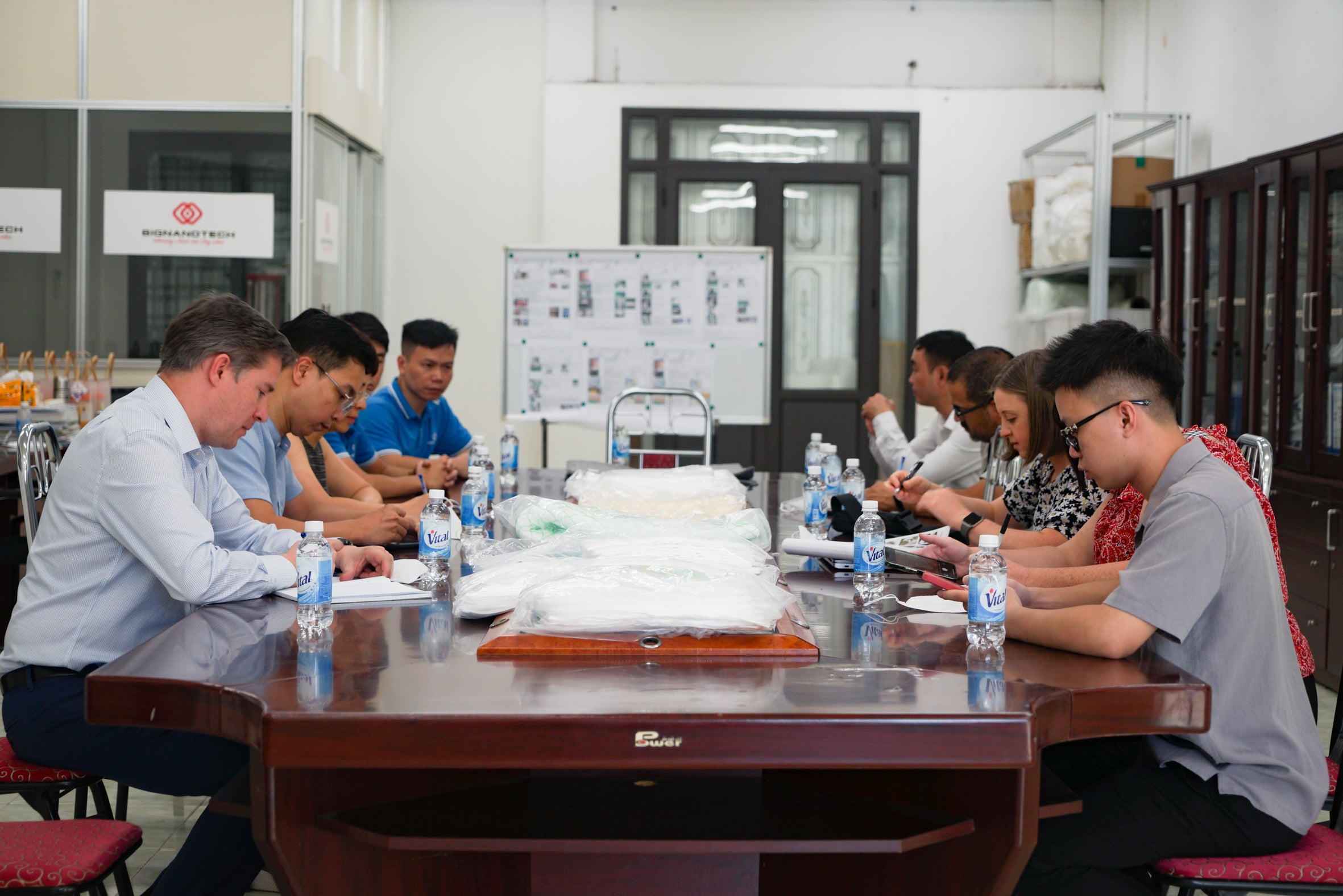 A group of people in business casual dress sit around a long table and examine a pile of white, cloth-like items.