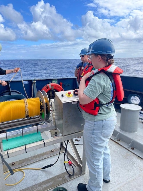 woman in life vest stands at control panel. Beyond her is open sea.