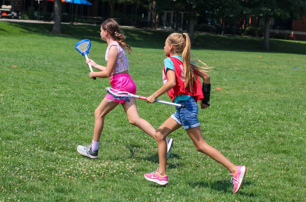 two young girls run across a field holding lacrosse sticks