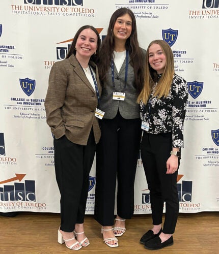 Three persons pose in front of an event backdrop 