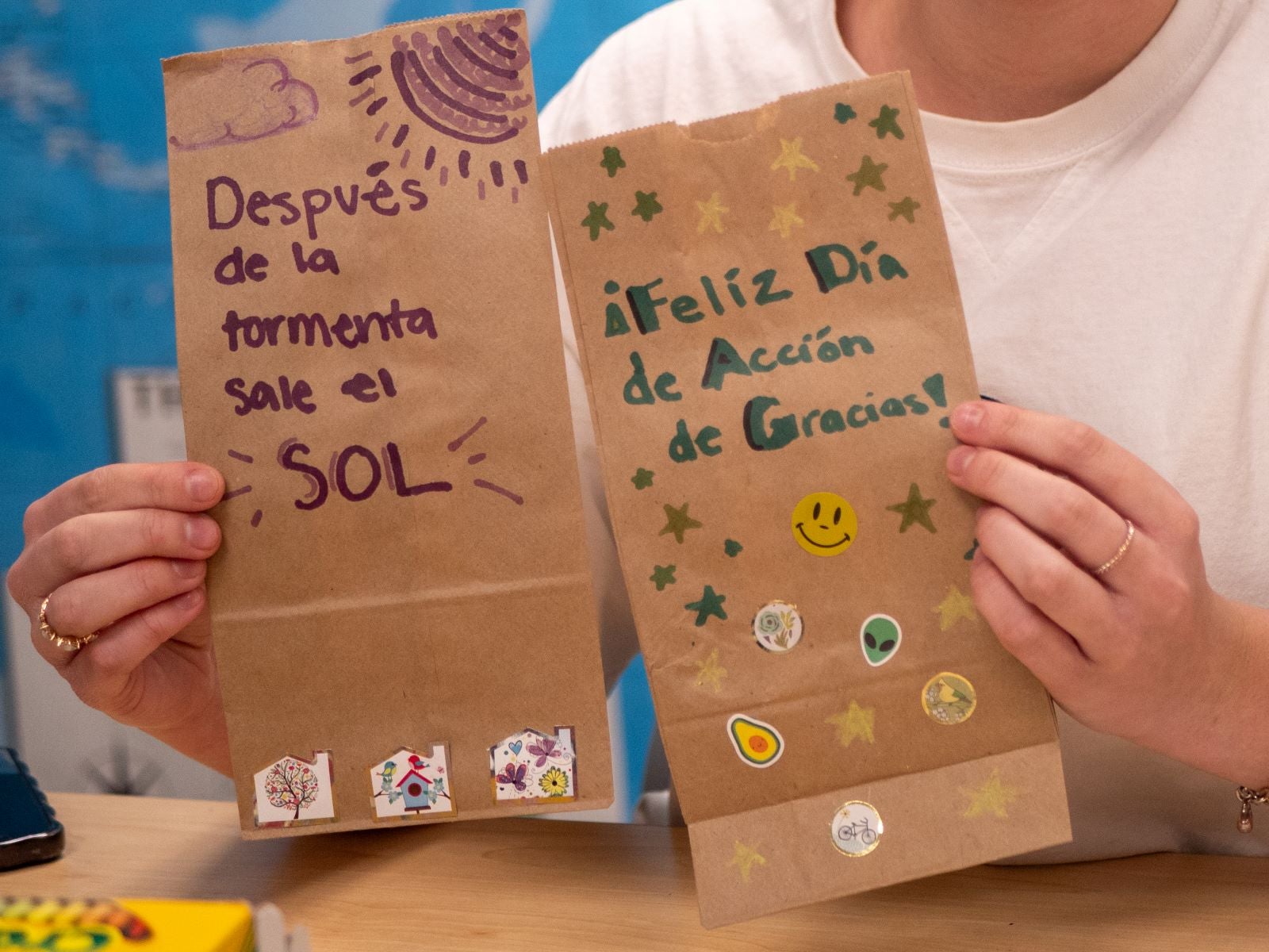 A person holds tow paper bags decorated with sayings in Spanish: "After the storm, the sun comes out." and "Happy Thanksgiving Day" 