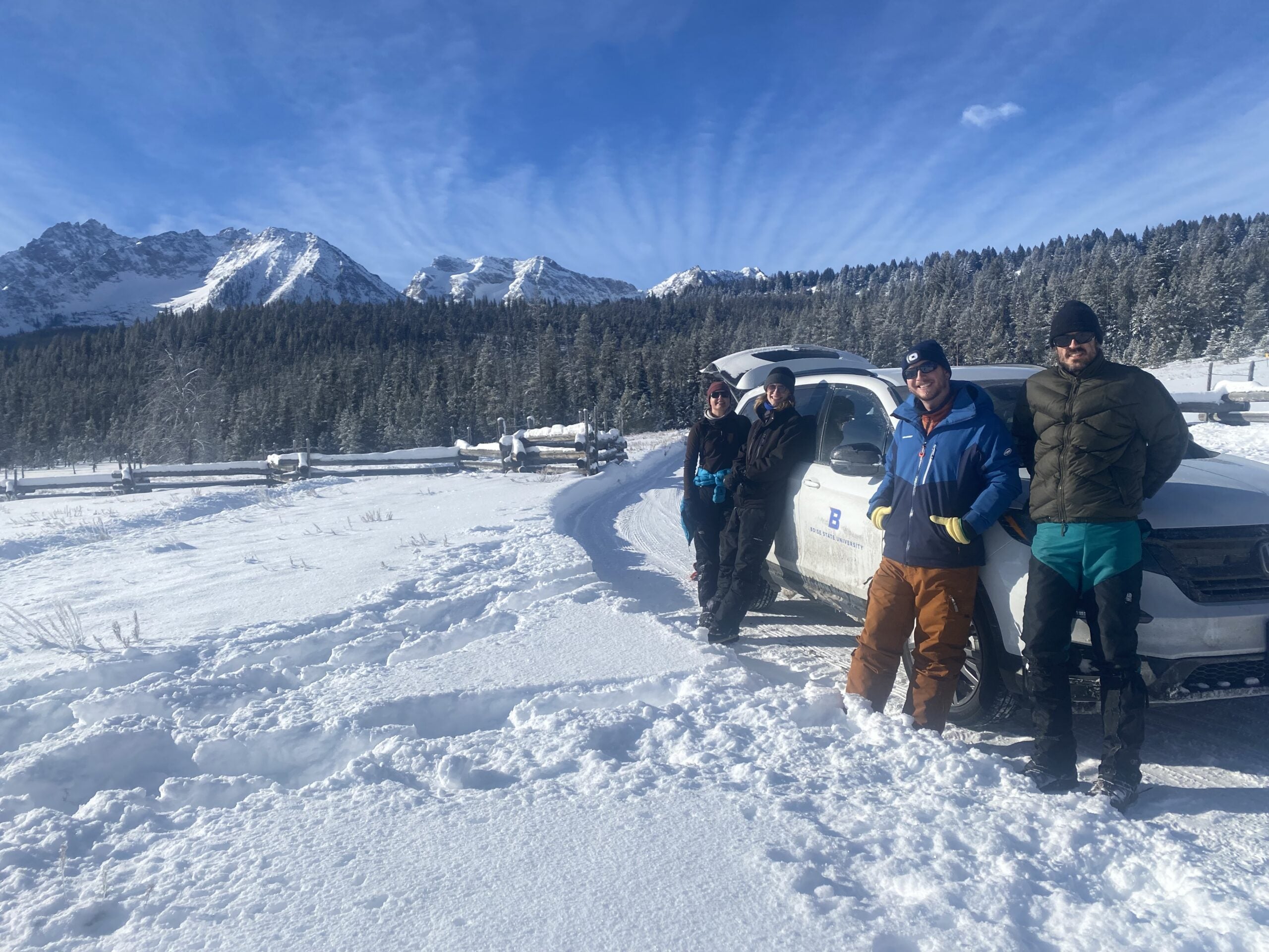 Persons stand in a snow field next to a white SUV