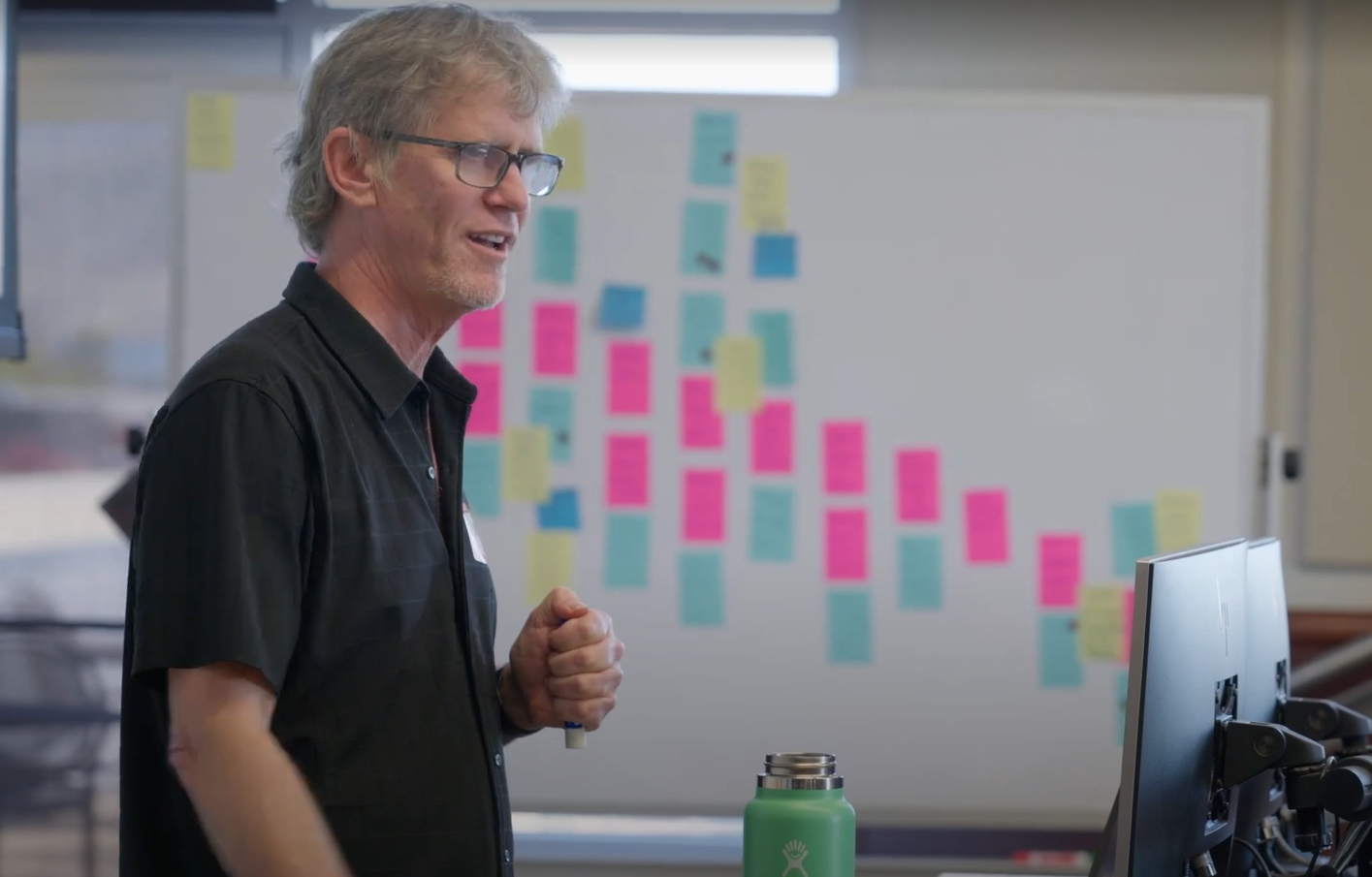 A man presents to a group with a whiteboard in the background