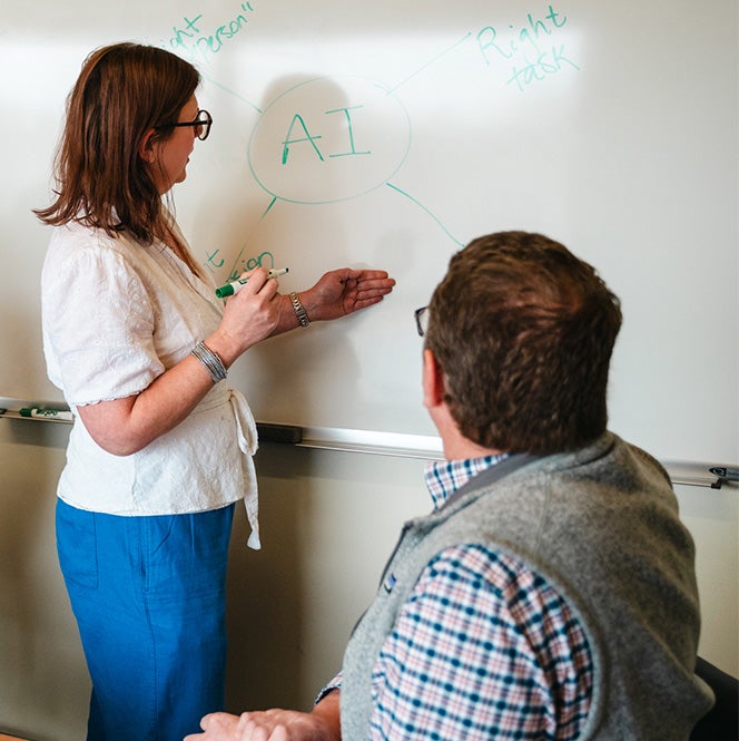 A teacher stands and points at a white board that says "AI" while another person with their back to the camera looks on.