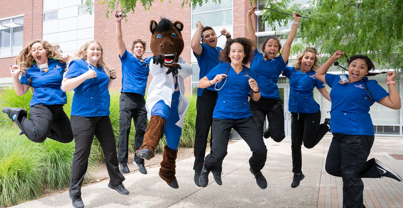 Students in scrubs jump with Buster Bronco in front of the Norco building