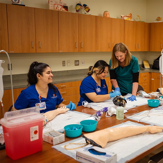 Two nursing students practice placing IVs while a nurse faculty assists.