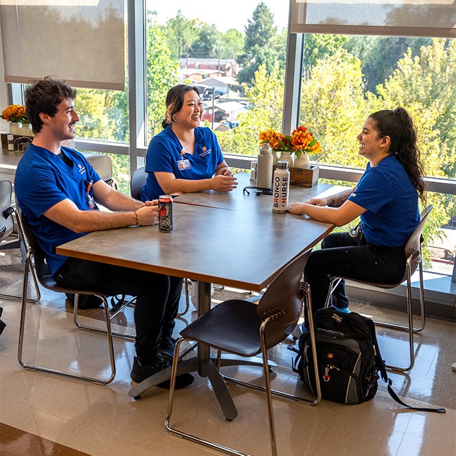 Three nurses talk and laugh together while sitting at a table by a wall of windows.