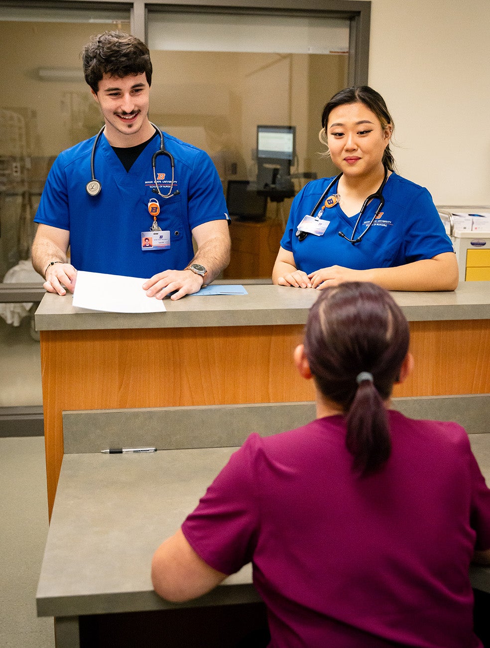 Two nurses stand at a Nurses Station and talk to a nurse behind the desk.