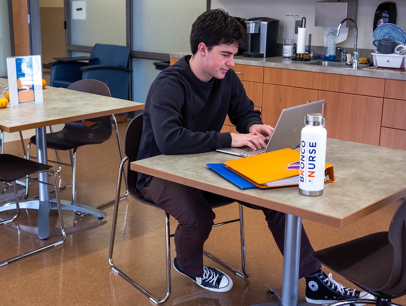 A man studies on a laptop at a break room table with a sink in the background and a water bottle that reads "Bronco Nurse" in the foreground.