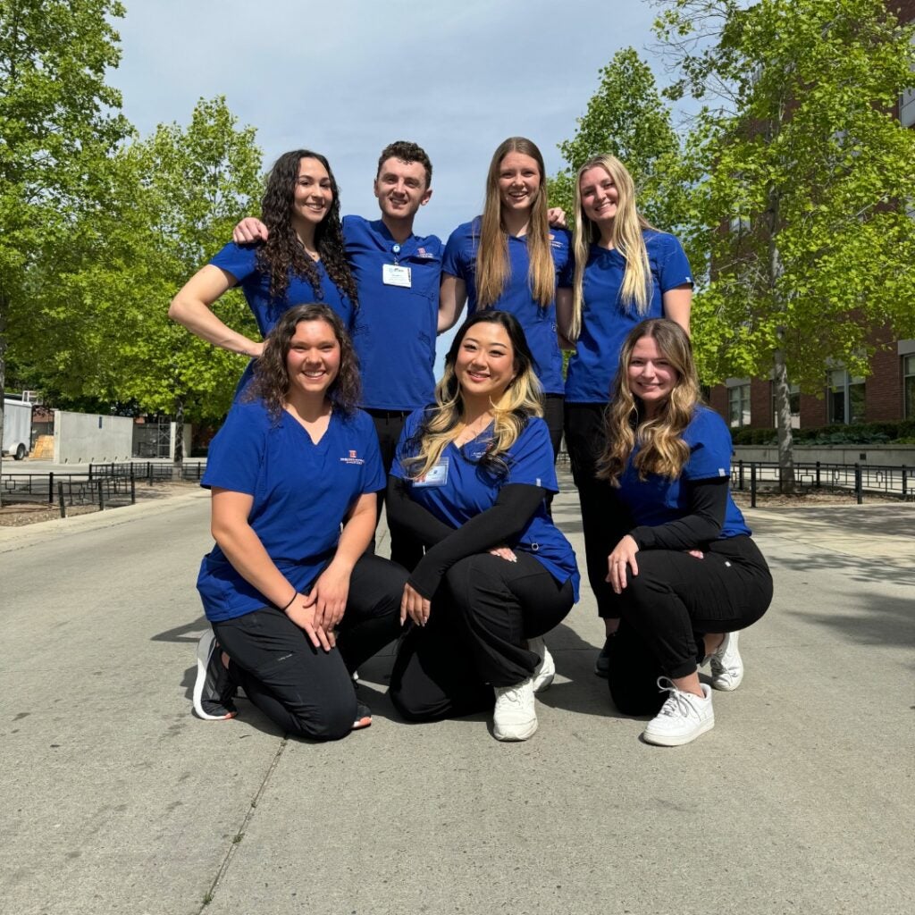 Seven students in blue scrub tops group together outside and smile.