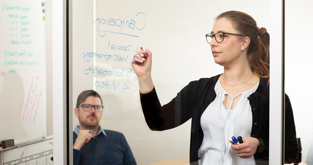 Man takes notes as woman writes marketing strategy on whiteboard