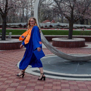 Graduate Caitlin Vasko in a blue robe with an orange sash walks by a large circular statue