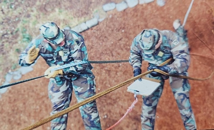 Two men in military uniforms rappel on a rock face