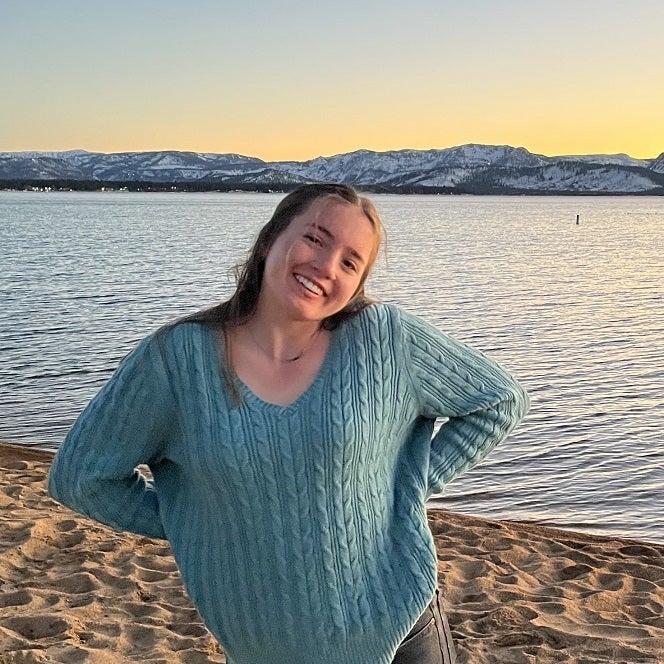 Emma standing on the shore of a lake, smiling with her hands on her hips