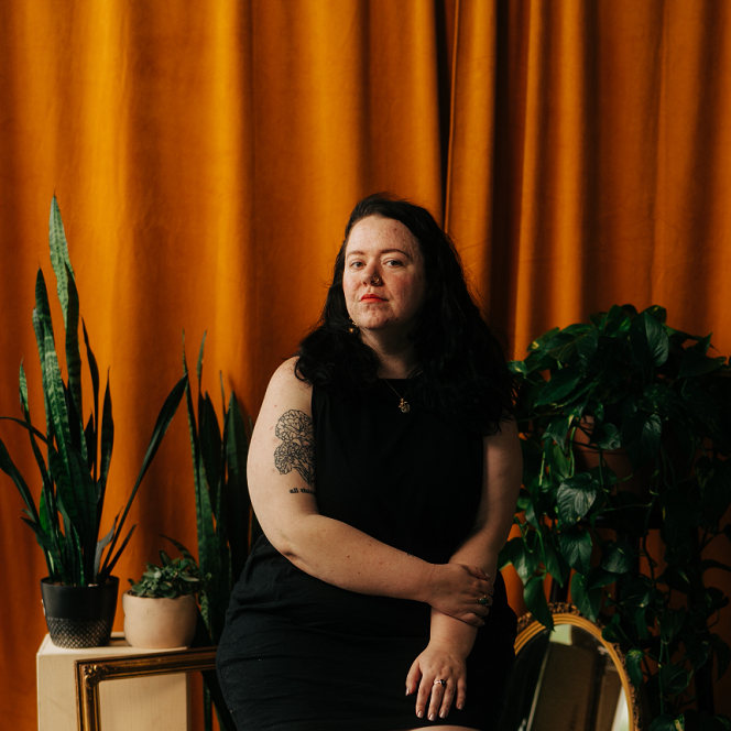 Lyd, a white person with dark brown hair in a black dress, is in front of an orange background and various plants.