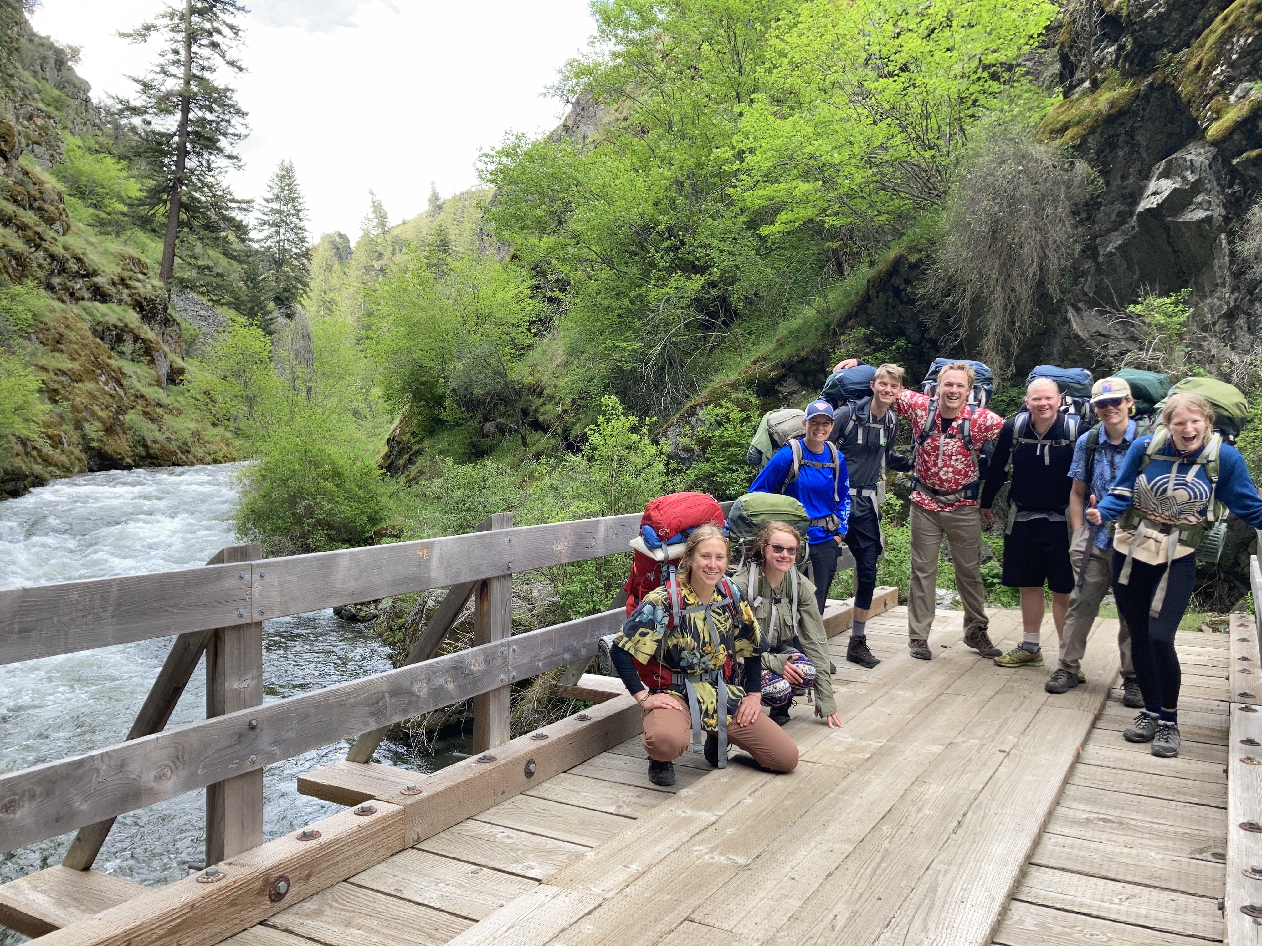 students stand on a bridge over a river wearing backpacking gear