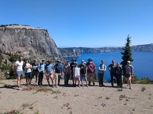 The image shows 15 hikers standing on a cliffside, overlooking a deep blue lake with rugged terrain and a clear sky.