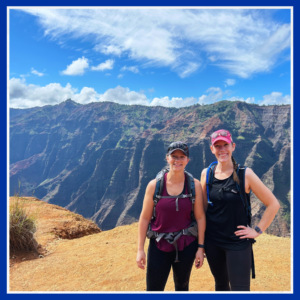 Cassie stands with her college roommate while hiking Waimea Canyon, Kauai. A large canyon and mountains are in the background.