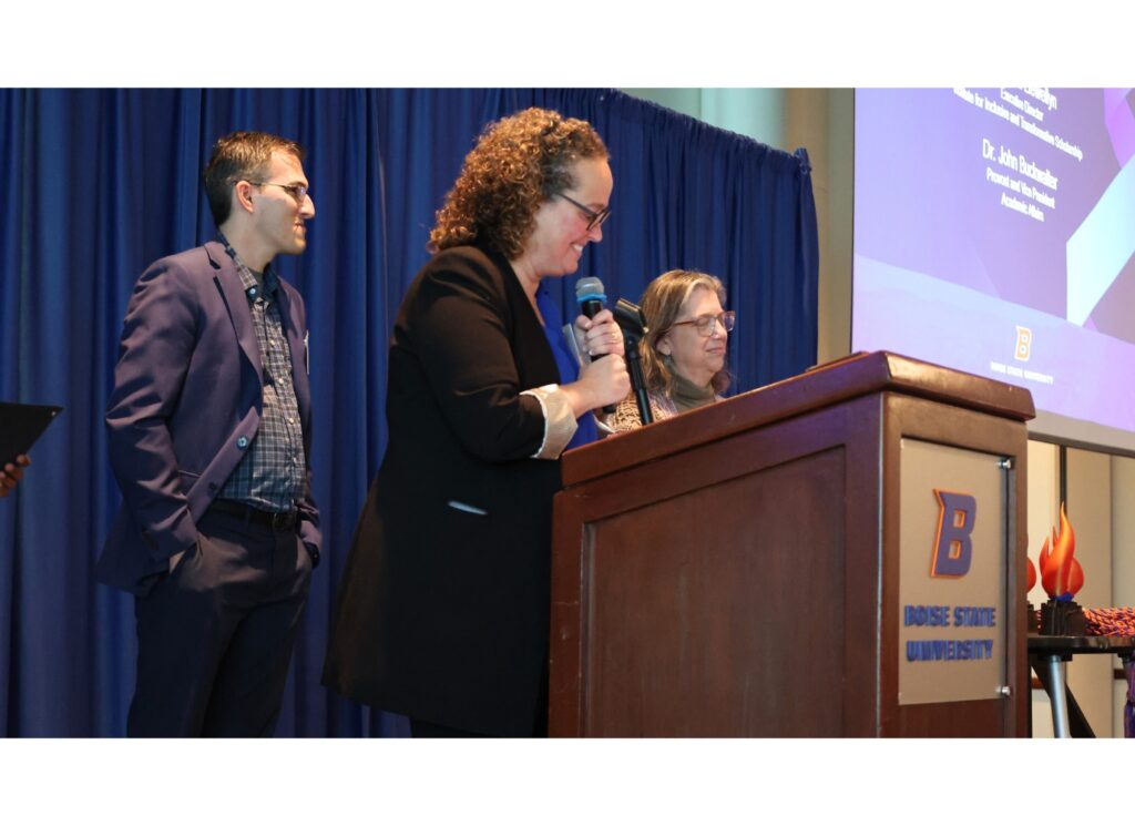 woman speaking at podium with two colleagues on stage behind her