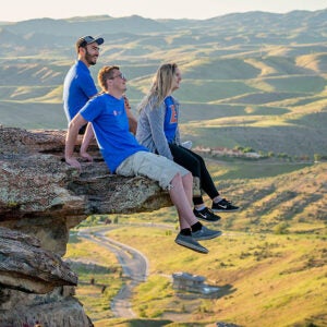 three students sitting on an edge of a rock and viewing the scene