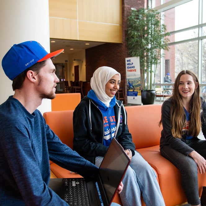 Three students, one in a baseball cap, one in a headscarf, one with long brown hair, sit on an orange couch and smile, looking at each other, as if in conversation