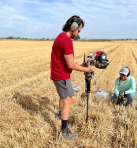 Man holding a soil auger in a wheatfield to drill into the ground.