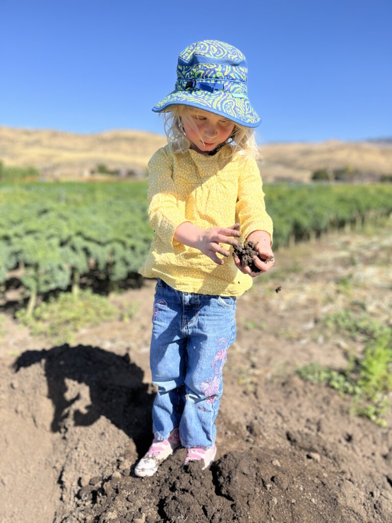 A young child wearing a bucket hat stands in a field with soil in her hand