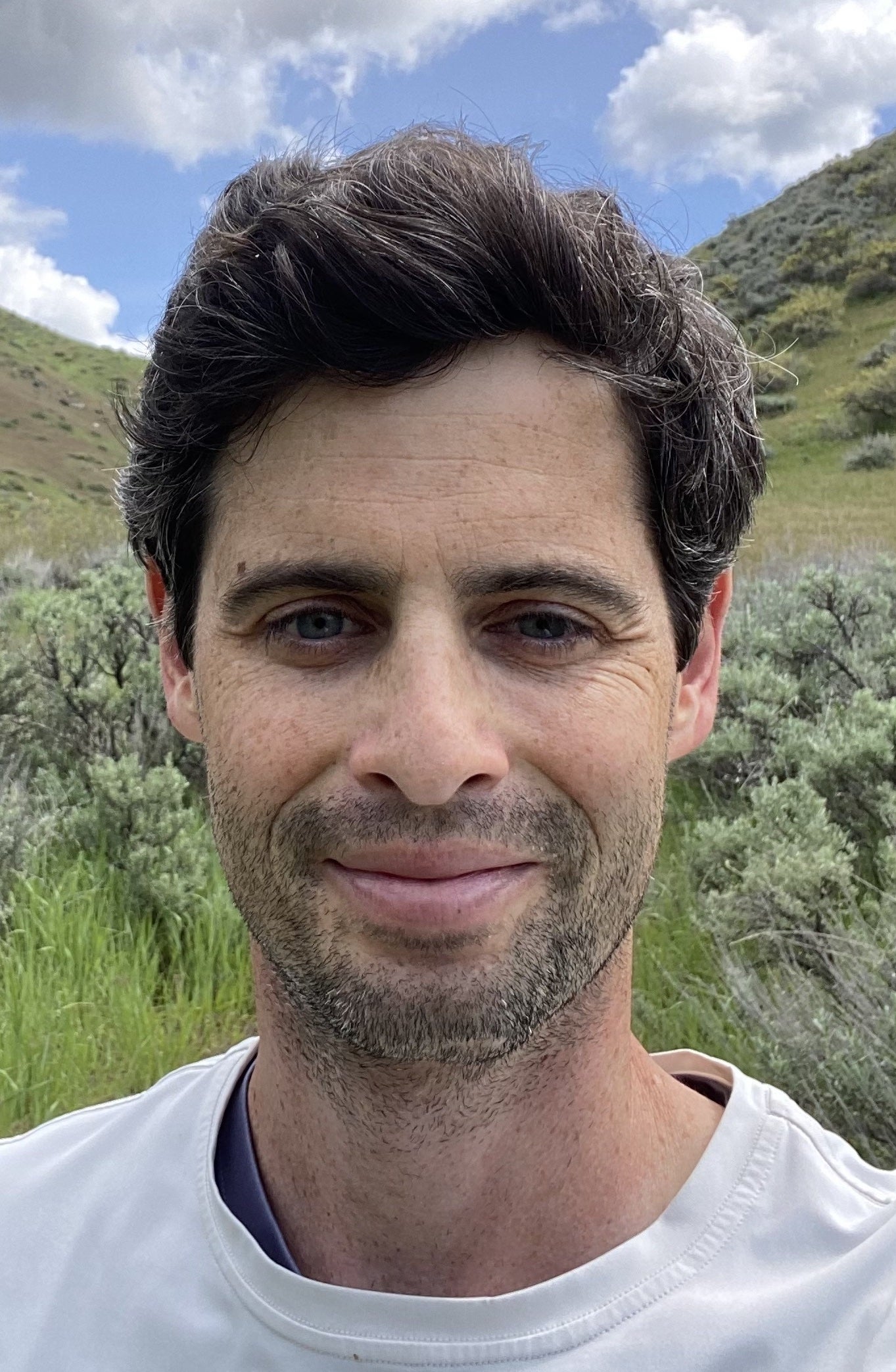 A man with light skin and stubble is standing in front of a green landscape. He has short brown hair and is smiling.
