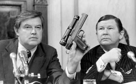 Photo of Frank Church in congressional hearing. Holding gun. 