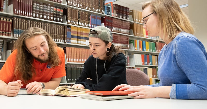 three students review a book at the library