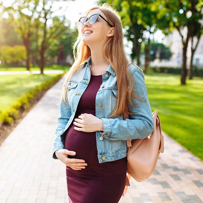 Pregnant person looking up at the sky with hands on belly against a campus sidewalk surrounded by grass and trees.