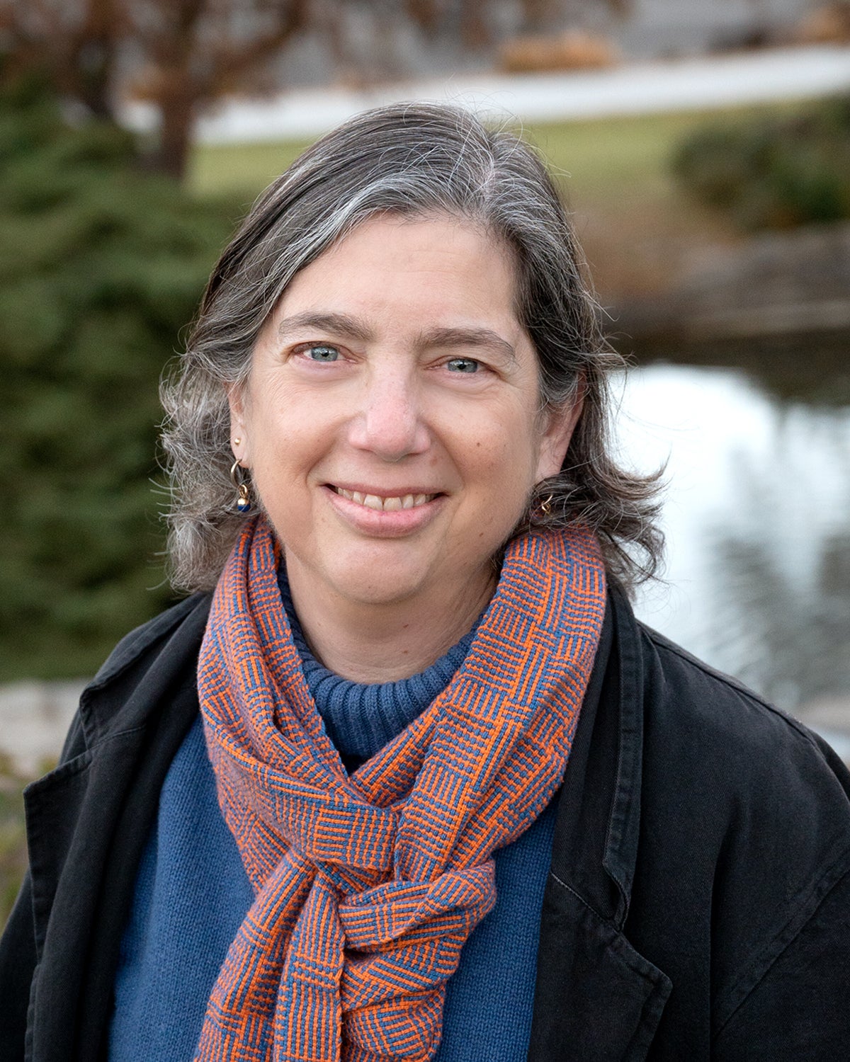 Profile picture of Donna Llewellyn wearing a blue and orange scarf, a blue turtleneck and a black blazer with trees and a pond in the background.