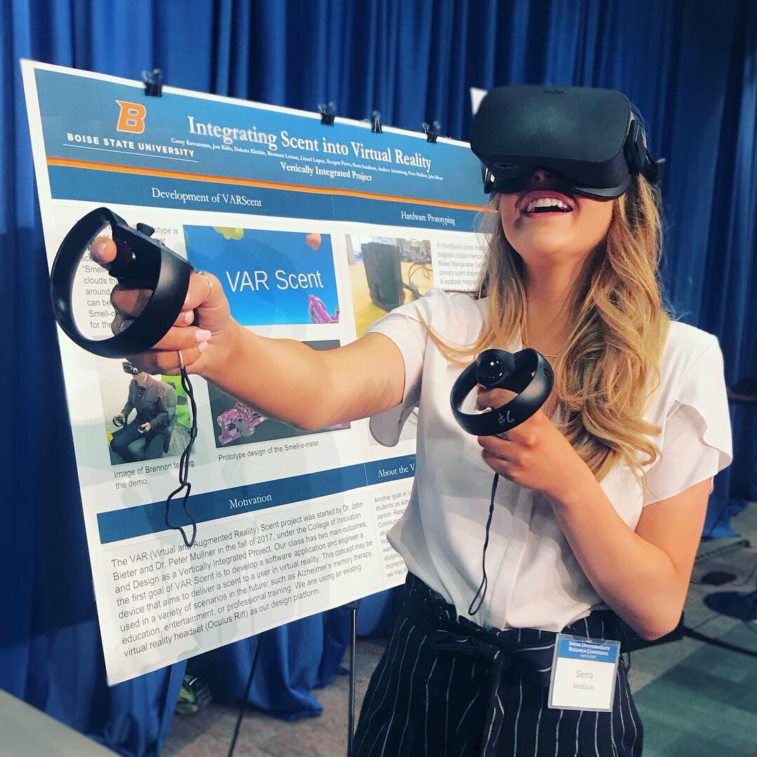 Student stands in front of academic poster at a poster conference. She is wearing a VR headset and is using hand controls.