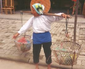 Elderly Asian woman carrying items in baskets suspended from opposite ends of a long stick slung over her shoulder; photograph