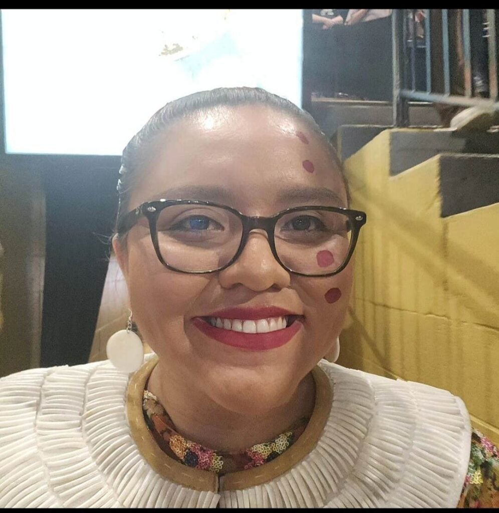 The image shows a smiling Veronica Yellowhair, graduate of the Department of Writing Studies BA in Writing, Rhetoric, and Technical Communication. She is wearing glasses, earrings, and a white outfit with decorative patterns. She has red circular markings on her face, as part of a cultural celebration. The background includes a staircase with yellow walls and a brightly lit screen.